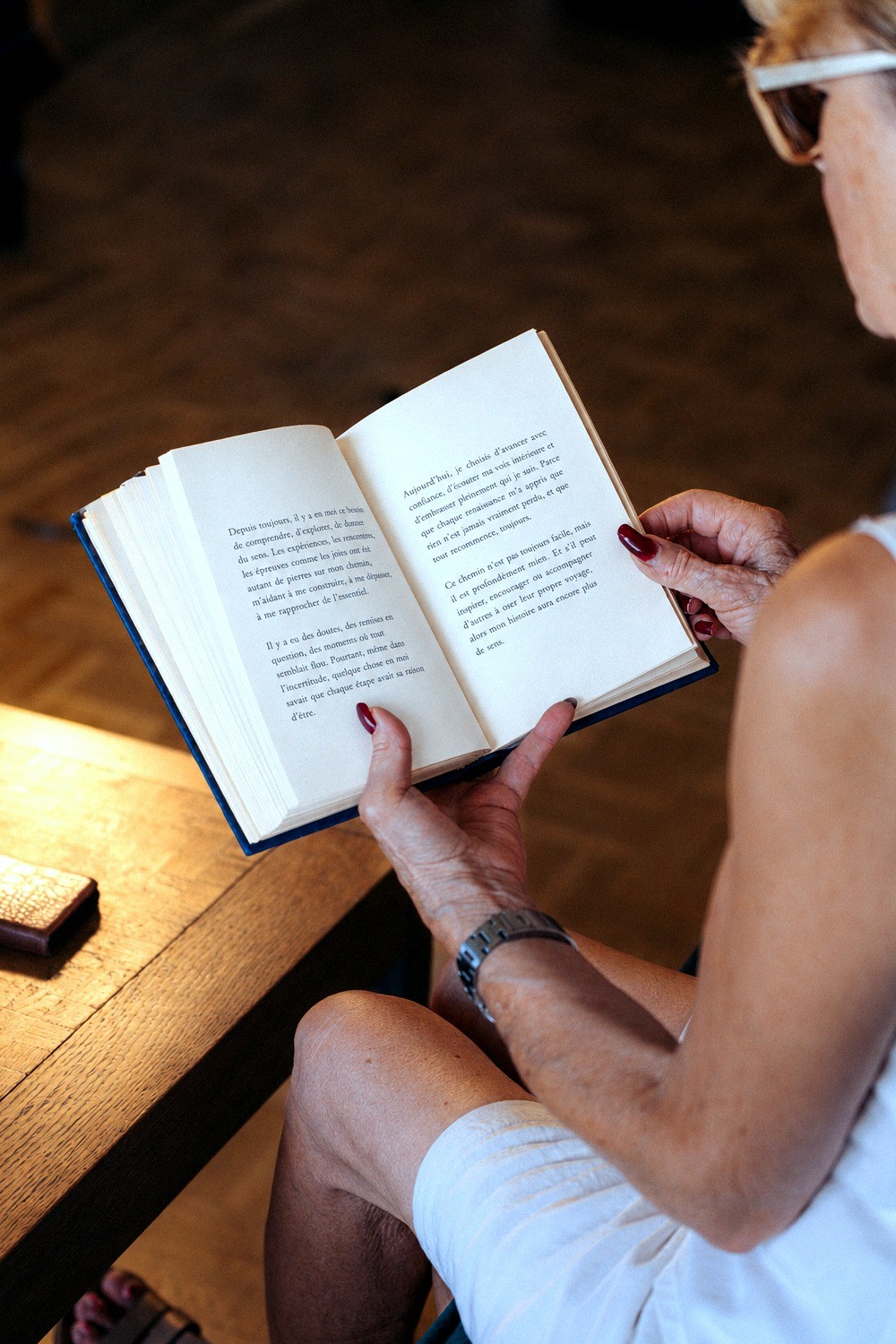 Une femme âgée lit un livre relié, assise près d'une table en bois.