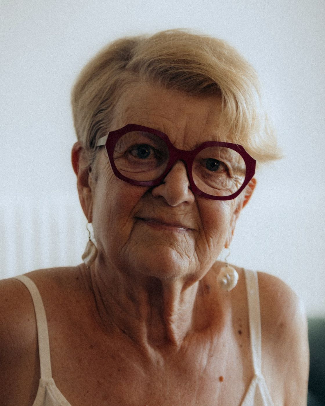 Une femme âgée aux lunettes rouges, chez elle, souriante.